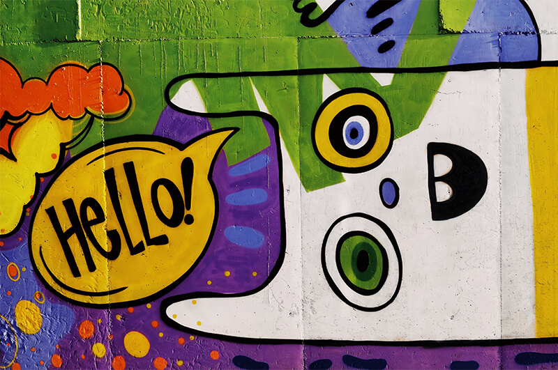 Auf dem Foto ist eine bunte Grafitti zu sehen: Ein auf der Seite liegendes Wesen mit Sprechblase, in der „Hello!” steht.